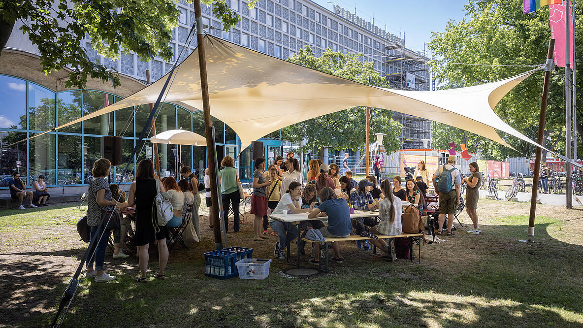 Unter einem weißen Sonnensegel vor dem Haupteingang der AGB ist eine große Gruppe von Kindern und Erwachsenen versammelt. Manche sitzen an einem Biertisch und malen, andere unterhalten sich im Stehen. Es ist ein sonniger Sommertag mit blauem Himmel. 