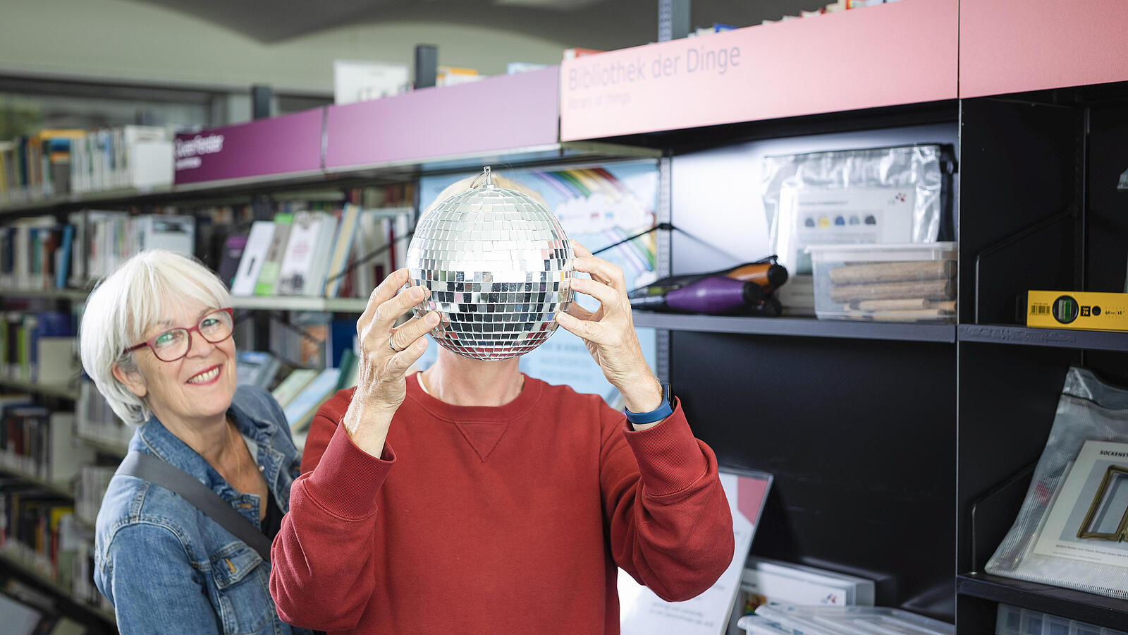 Ältere Frau steht mit älterem Mann, der sich Discokugel vors Gesicht hält, in Bibliothek