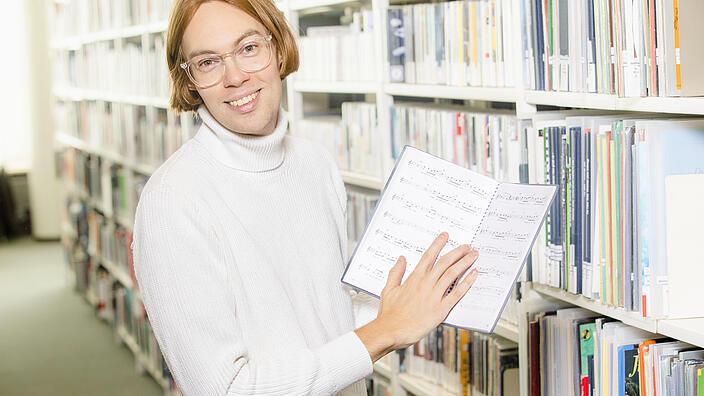 Young man with glasses stands in front of shelf in music library and holds open music book in hand