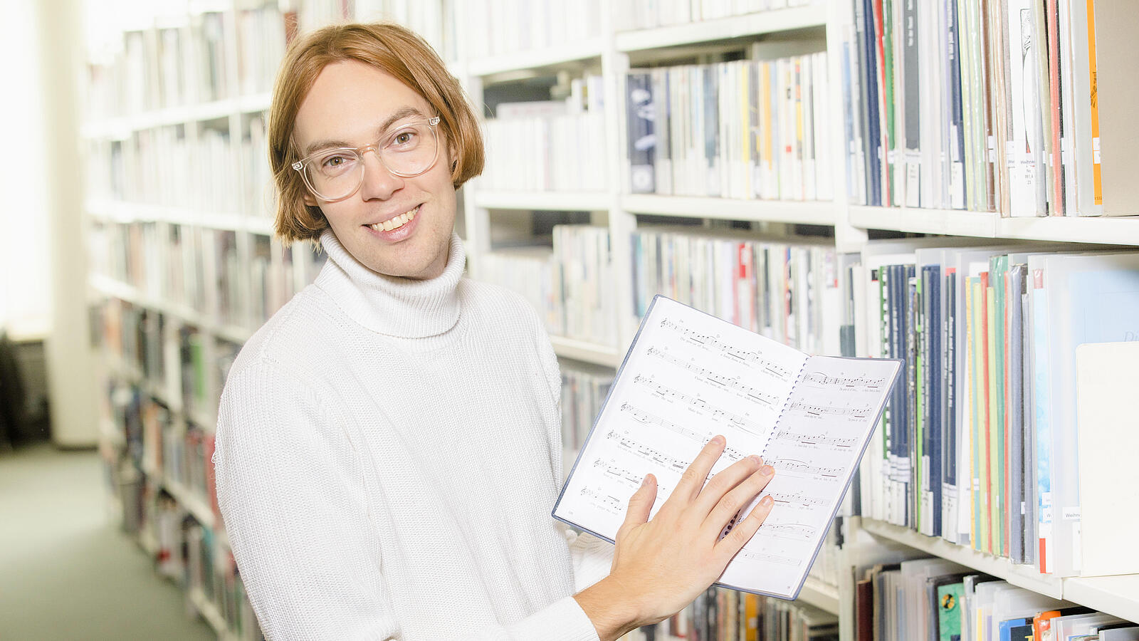 Young man with glasses stands in front of shelf in music library and holds open music book in hand