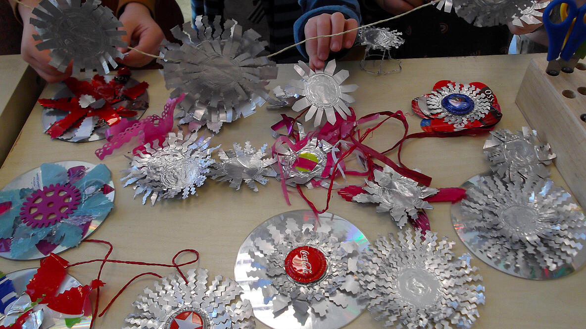 mehrere Kinderhände basteln Objekte aus CD´s, Alufolie, Kronenkorken und anderen recycelten Materialien