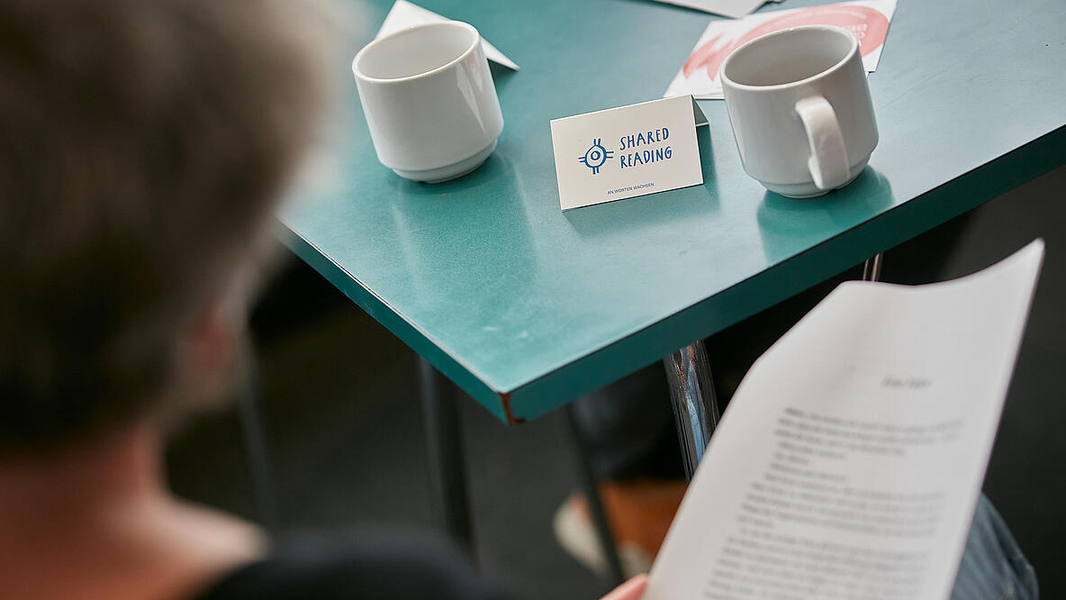 Eine Frau sitzt an einem grünen Tisch, in der Hand hat sie einen Text auf einem Stück Papier. Auf dem Tisch stehen Kaffeetassen und ein Schild, auf dem Shared Reading steht.