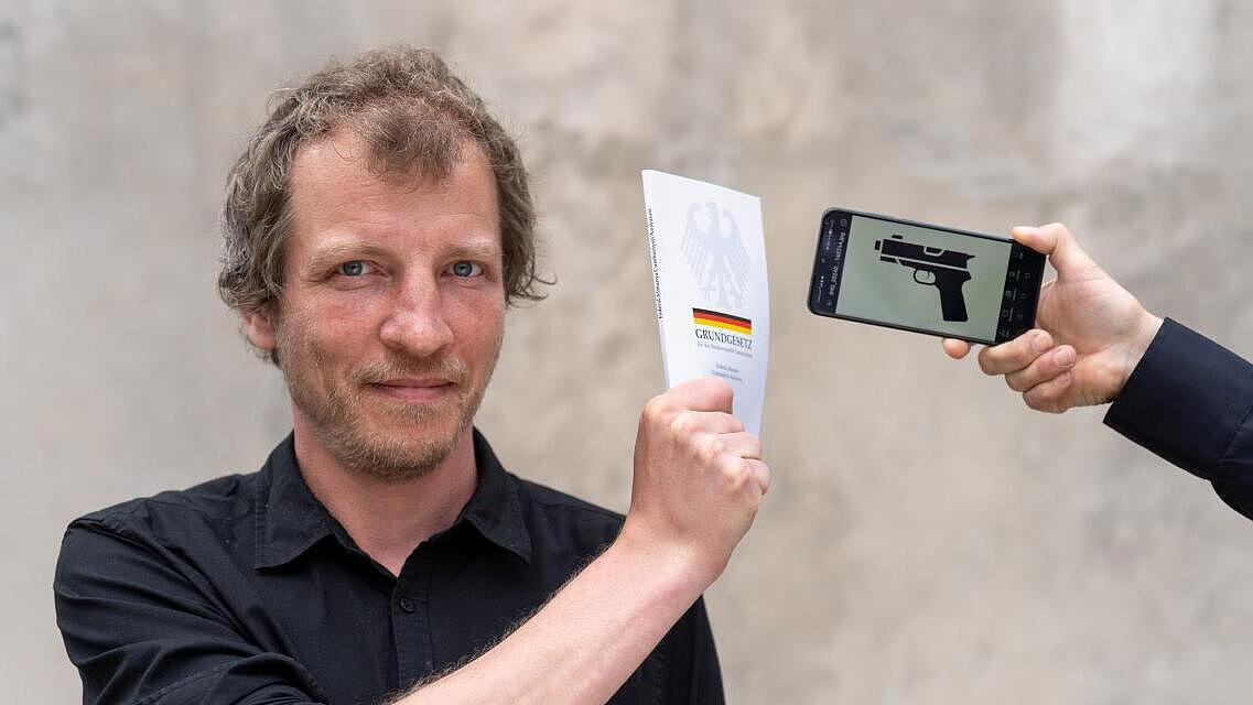 Foto einer Person, die das Grundgesetz in der Hand hochhält und damit ein ihm entgegengestrecktes Smartphone abwehrt, auf dem eine Pistole abgebildet ist.