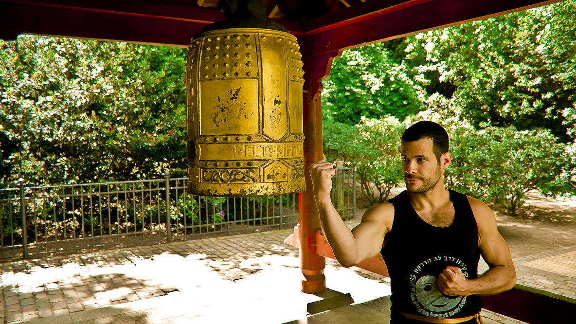 muskulöser Mann steht unter Pavillion in Natur vor einer goldenen Glocke in einer Kung Fu Pose