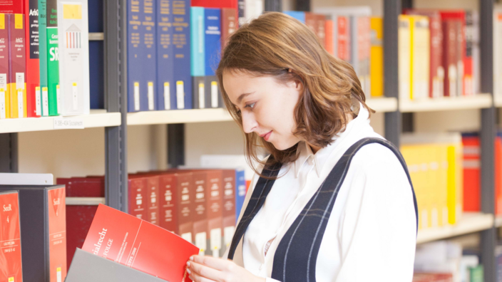 Junge Frau in Hosenanzug schaut sich ein juristisches Buch in der Bibliothek vor einem Regal an