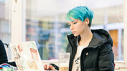 Junge Frau mit blauen Haaren steht in der Bibliothek an ihrem Laptop