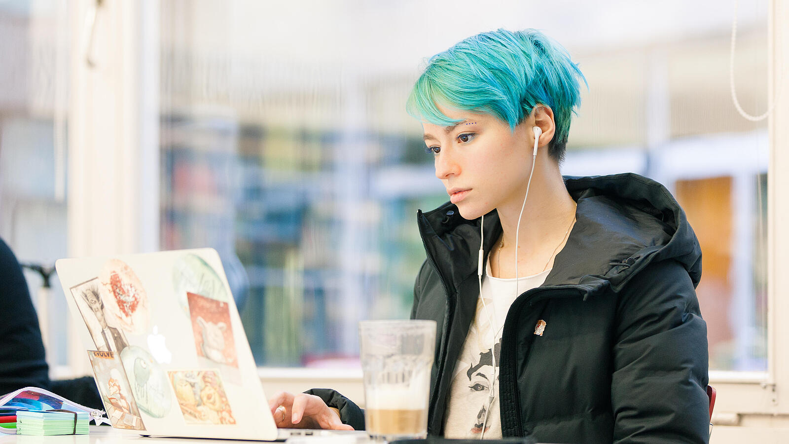 Junge Frau mit blauen Haaren steht in der Bibliothek an ihrem Laptop