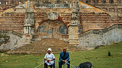 Foto zweier im Freien sitzender Personen vor einer Ruine, die linke Person hält altmodische Telefonhörer an einem Kabel und die rechte Person hält eine Ziege an einer Leine.