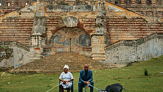 Foto zweier im Freien sitzender Personen vor einer Ruine, die linke Person hält altmodische Telefonhörer an einem Kabel und die rechte Person hält eine Ziege an einer Leine.