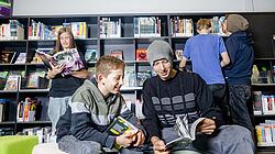 Junge und Jugendliche stehen oder sitzen in Zeitschriften-Ecke der Bibliothek mit Zeitschriften