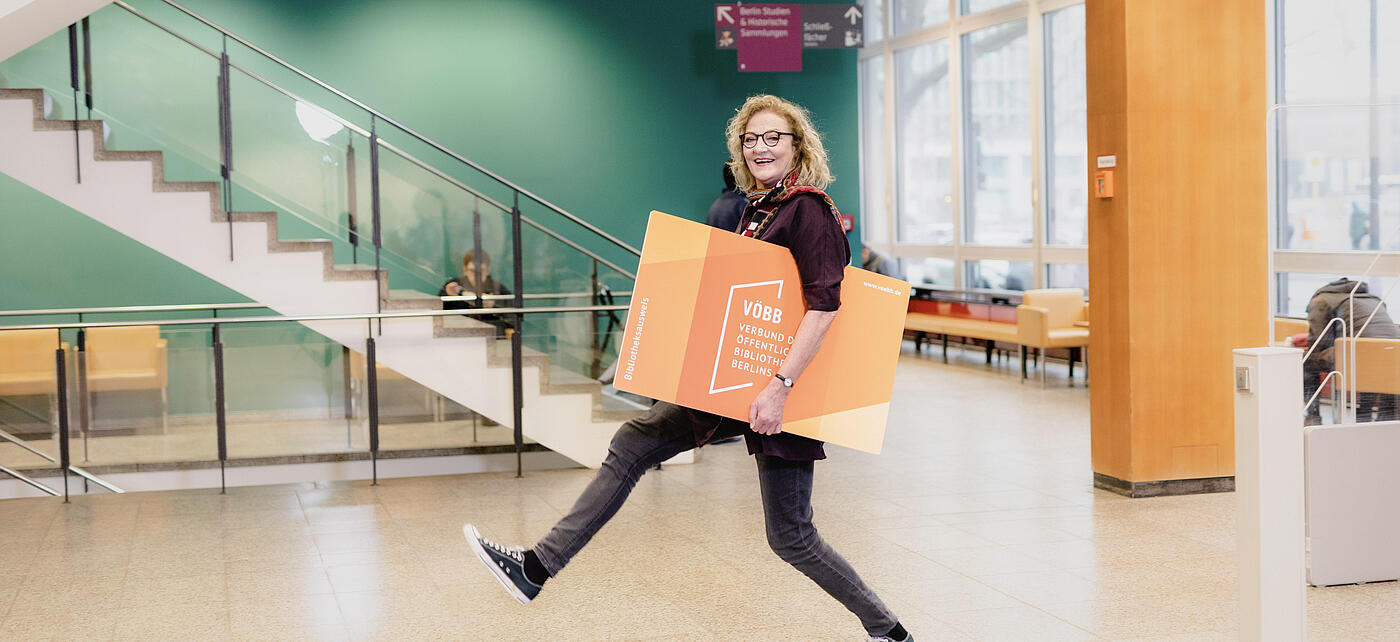 Woman with large library card under arm walks large stride through library foyer