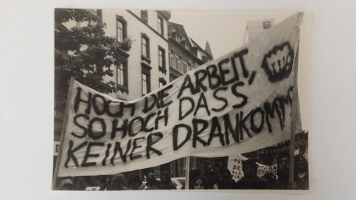 Schwarz-weiß-Bild eines Demonstrationsumzugs mit einem Banner mit der Aufschrift "Hoch die Arbeit, so hoch dass keiner drankommt"