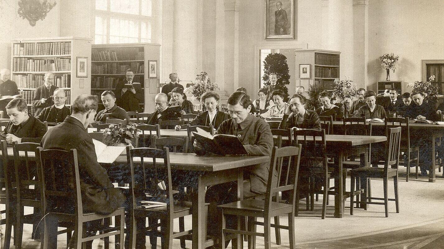 Historische Aufnahme von Menschen mit aufgeschlagenen Büchern an langen Holztischen in Bibliothek