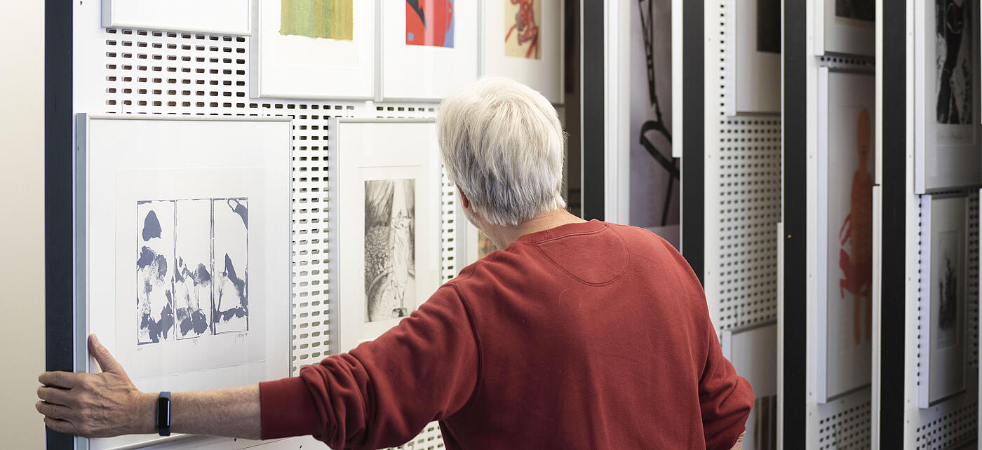 Älterer Herr in der Artothek in der AGB  hat das Auszugsregal herausgezogen und betrachtet die Kunstwerke