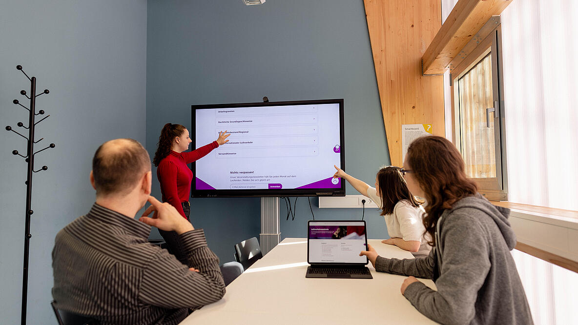 Personen sitzen um Tisch herum und schauen auf Bildschirm an der Wand.