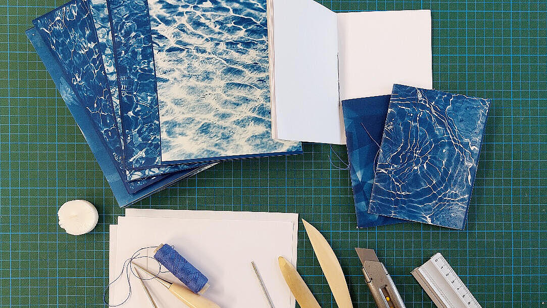 mehrere selbstgebundene Blaudrucke liegen zusammen mit Buchbindewerkzeugen wie beispielsweise Pfalzbeilen, Nadeln und einem Lineal auf einer Schneidematte ausgebreitet