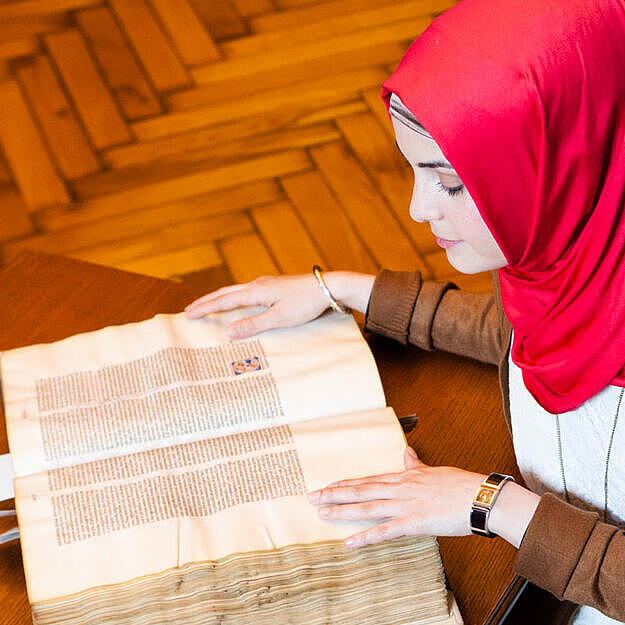 Young woman with hijab in historical reading room sitting bent over historical document