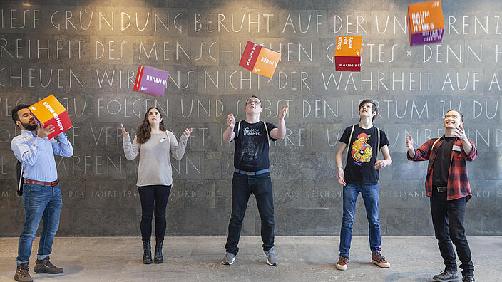 Auszubildende im Foyer der AGB werfen Pappwürfel mit der Aufschrift Raum für neues in die Luft
