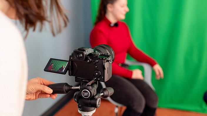 Foto einer Person, die eine Kamera bedient, im Hintergrund eine andere Person vor einem Greenscreen.
