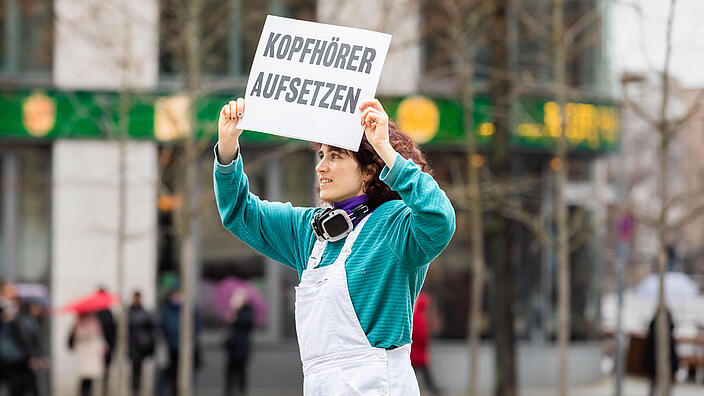 Eine Frau helt ein Schild mit der Aufschrift "Kopfhörer aufsetzen" 