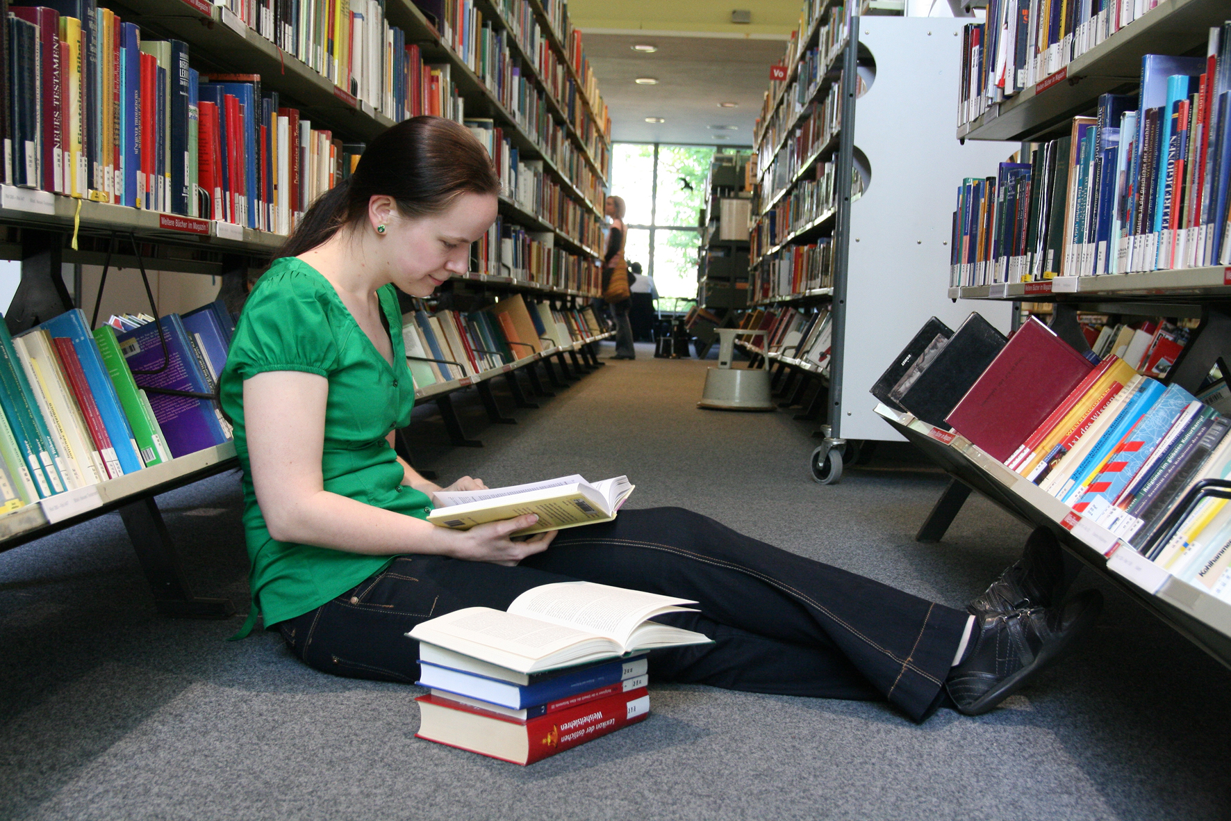 Nutzerin sitzt mit Bücherstapel zwischen den Regalen der AGB auf dem Boden