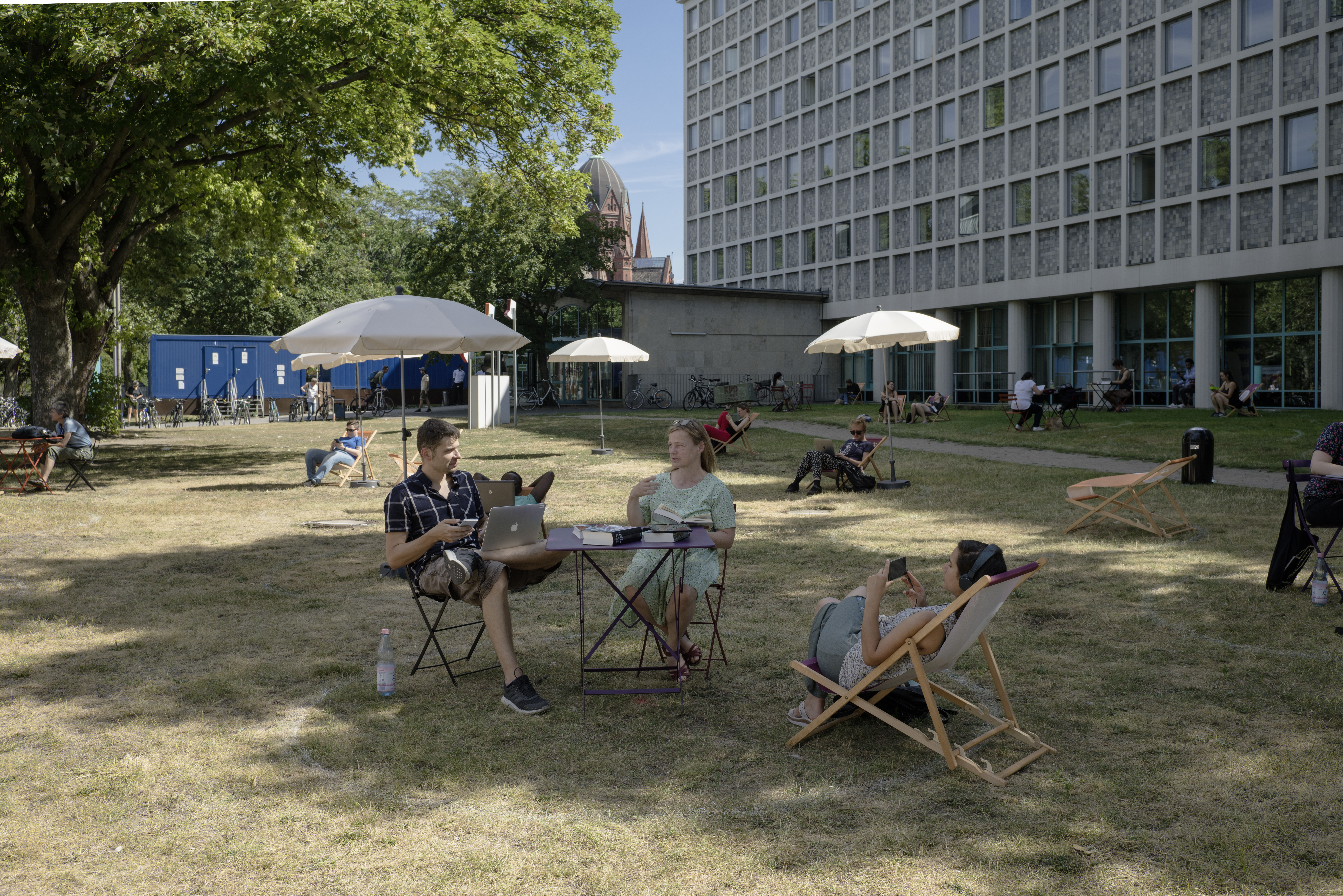 Menschen sitzen auf Gartenmöbeln auf Wiese vor AGB mit Sonnenschirmen im Hintergrund - die Frischluftbibliothek vor der AGB