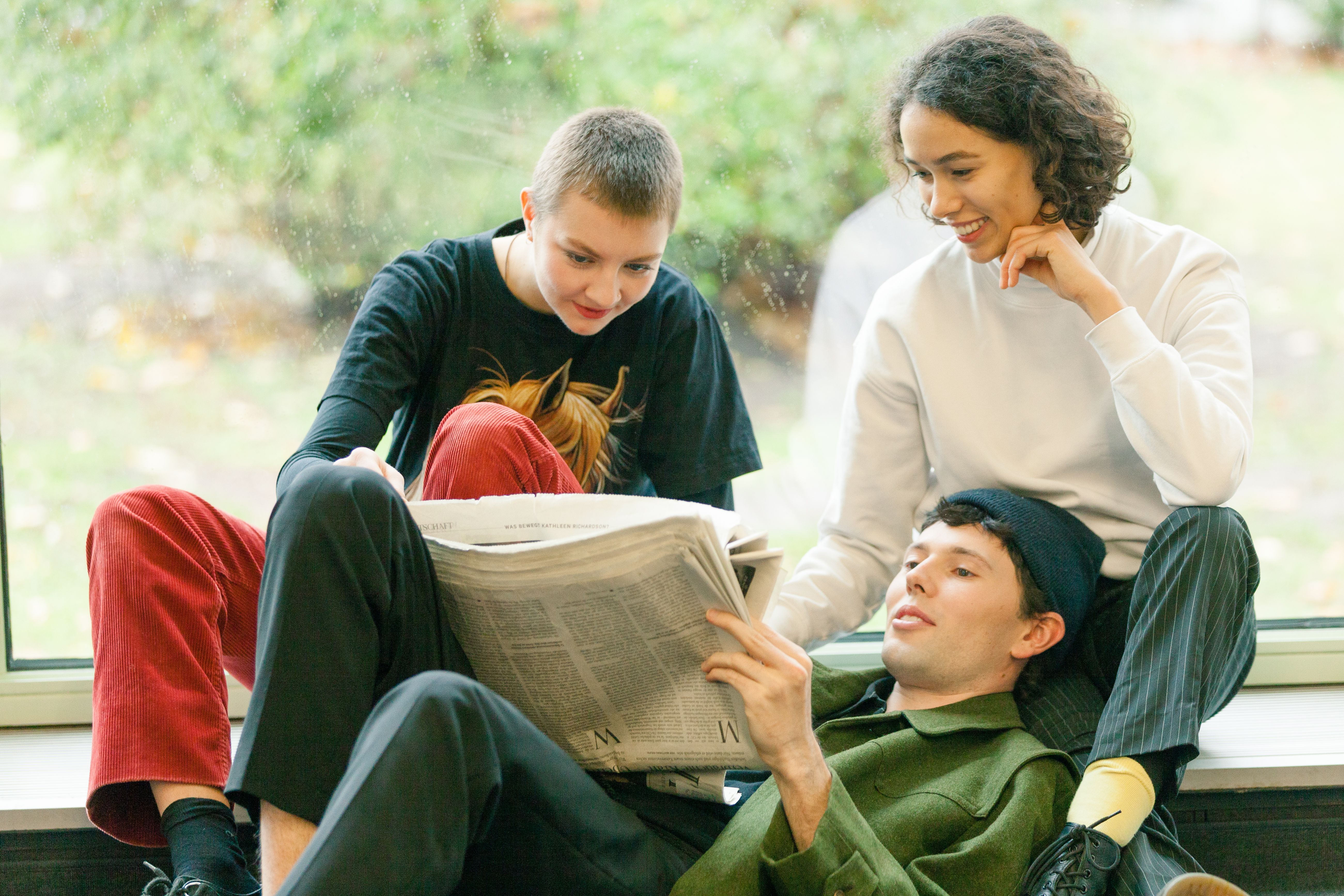Gruppe von 3 jungen Menschen schaut gemeinsam in eine Zeitung und sitzt dabei vor Fenster