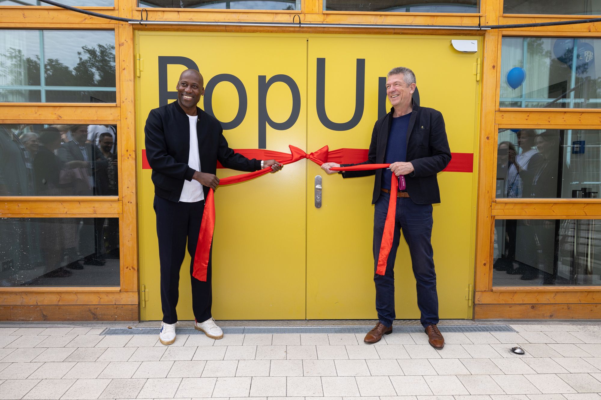 Foto von Kultursenator Joe Chialo und Generaldirektor der ZLB Volker Heller vor der Eingangstür zum PopUp-Gebäude. In den Händen halten sie eine große rote Schleife zur Eröffnung.