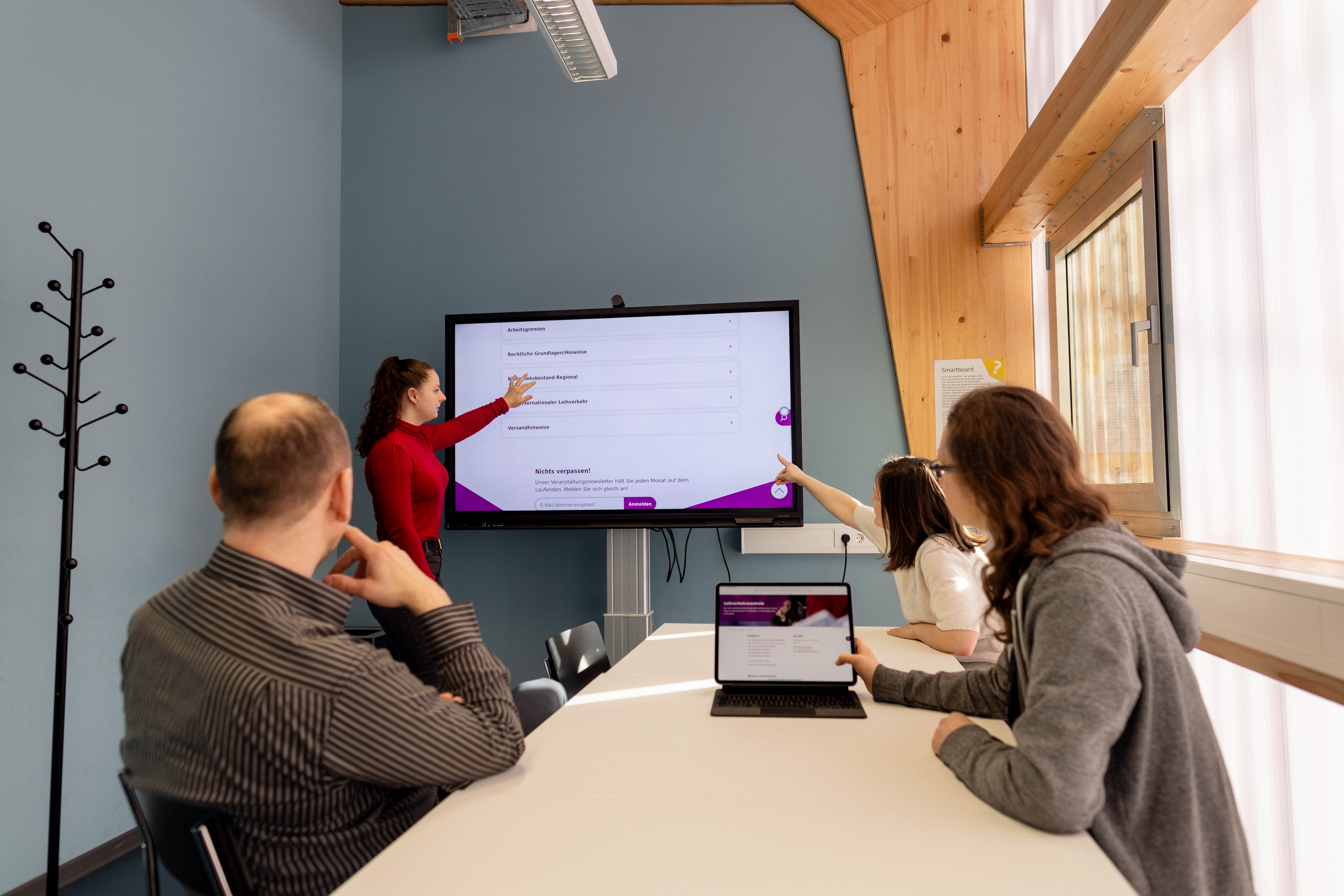 Personen sitzen um Tisch herum und schauen auf Bildschirm an der Wand.