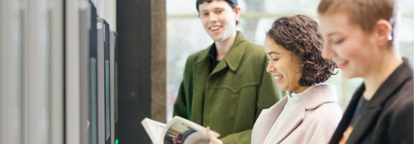 Three smiling persons in a room, two of them reading a book