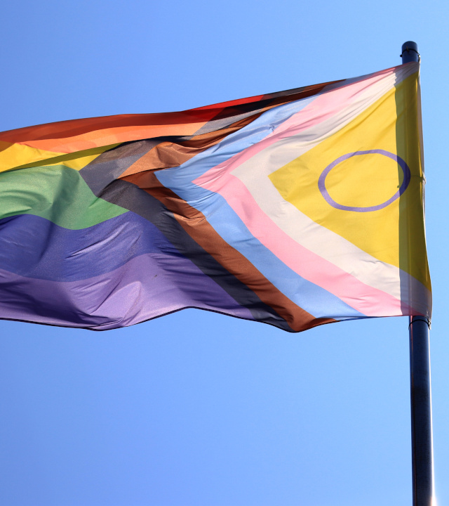 Eine Progress-Pride-Flagge weht an einem Fahnenmast vor einem klaren blauen Himmel.